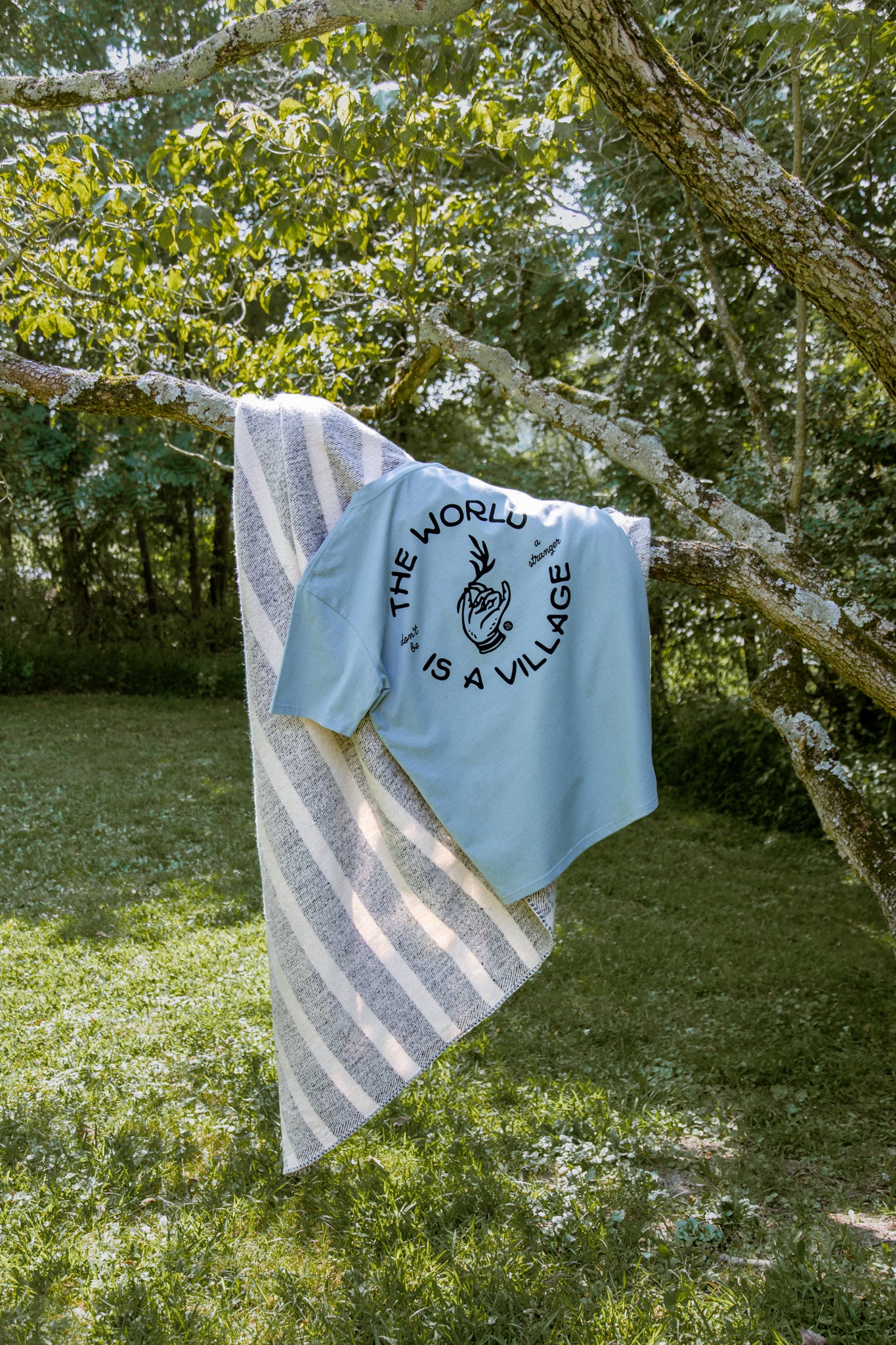 Light blue t-shirt with 'The World is a Village' text hanging on a tree branch with a striped towel.
